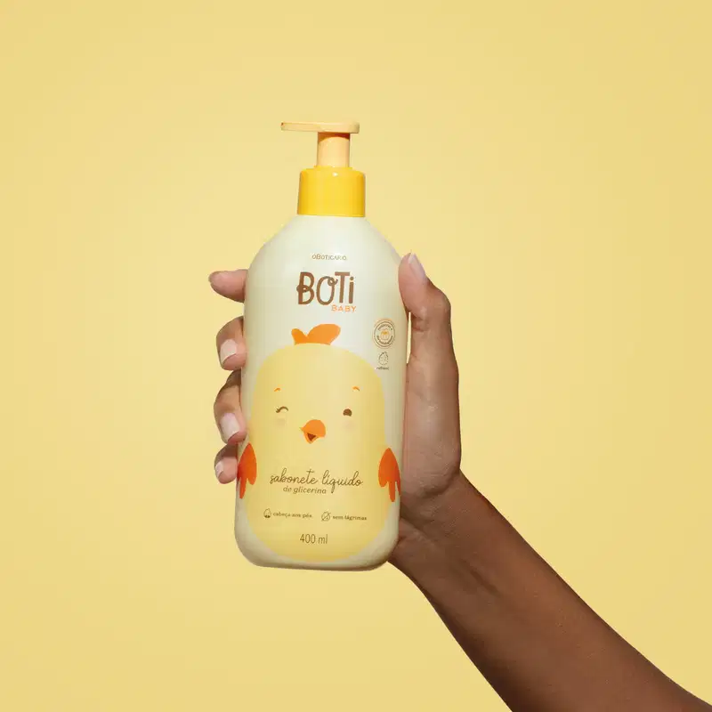 Mão segurando um frasco amarelo claro com ilustração de um passarinho amarelo com detalhes, tampa e bomba dosadora laranjas contendo sabonete líquido de glicerina da marca Boti Baby em um fundo alaranjado.