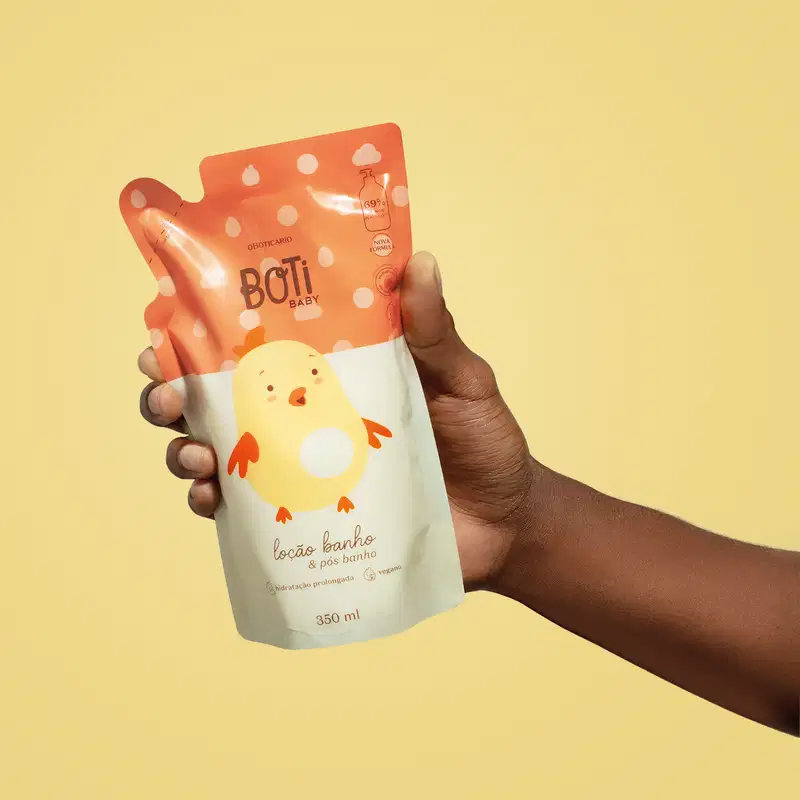 Mão femininas segurando um refil de loção hidratante corporal da marca Boti Baby em forma de sachê nas cores laranja e amarelo claro com ilustração de passarinho amarelo com detalhes laranjas. O fundo da imagem é laranja claro.