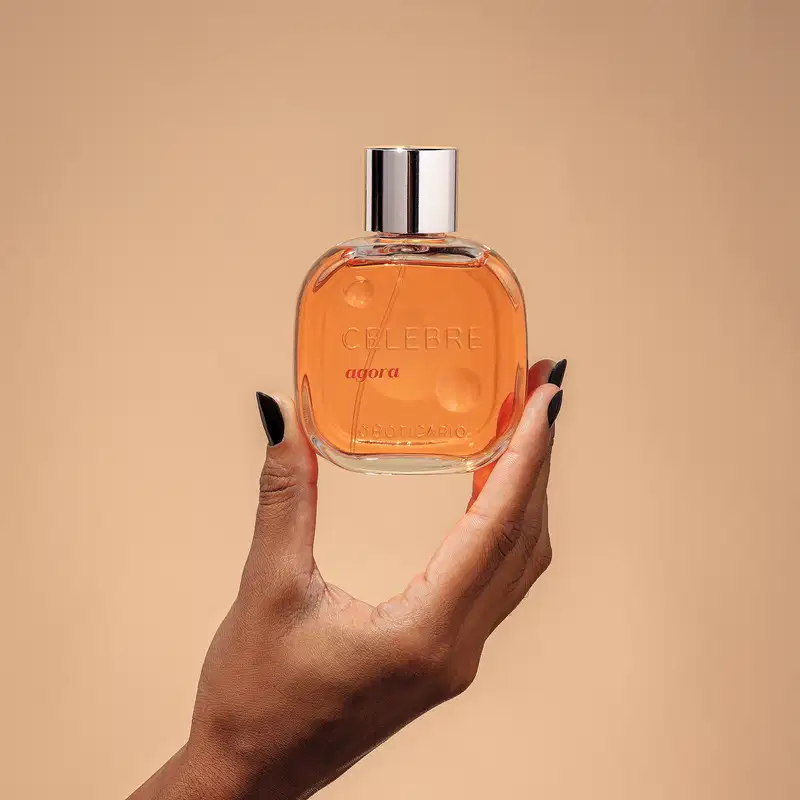 Uma mão feminina com unhas pintadas de preto segurando um frasco de vidro transparente com líquido laranja, tampa prateada e letras pretas e em relevo de desodorante colônia Celebre Agora Feminino em fundo em tons de laranja.