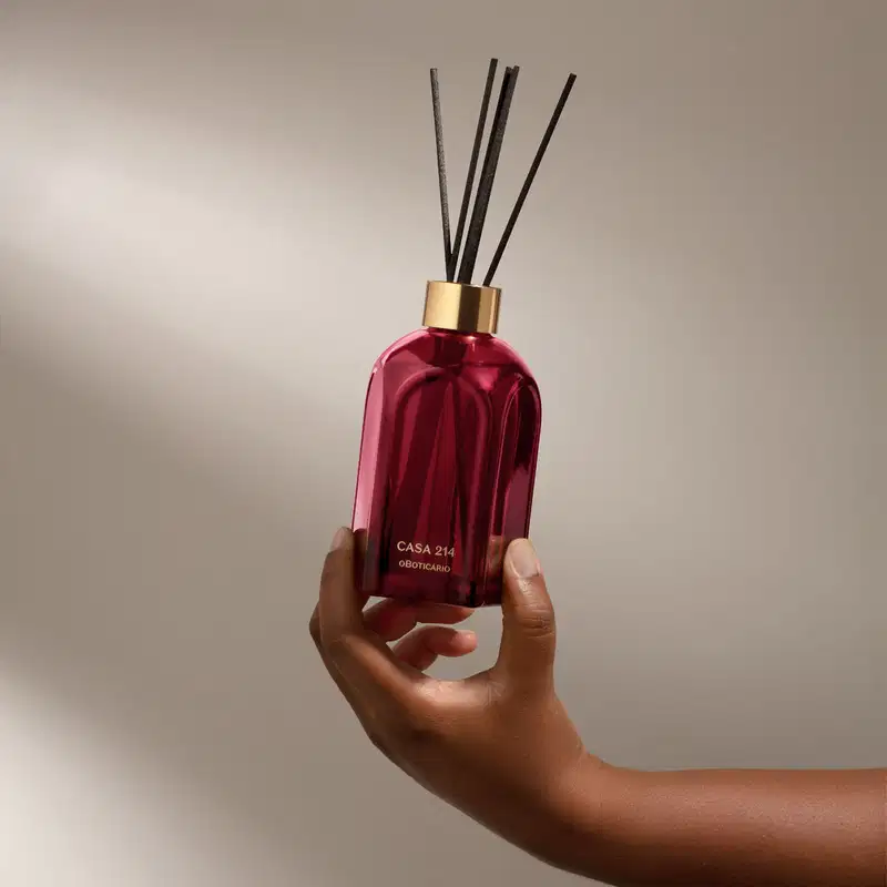 Mão segurando um Difusor de ambientes em vidro vermelho com varetas e letras douradas da marca Casa 214 Gardênia Imperial em fundo cinza com sombras.