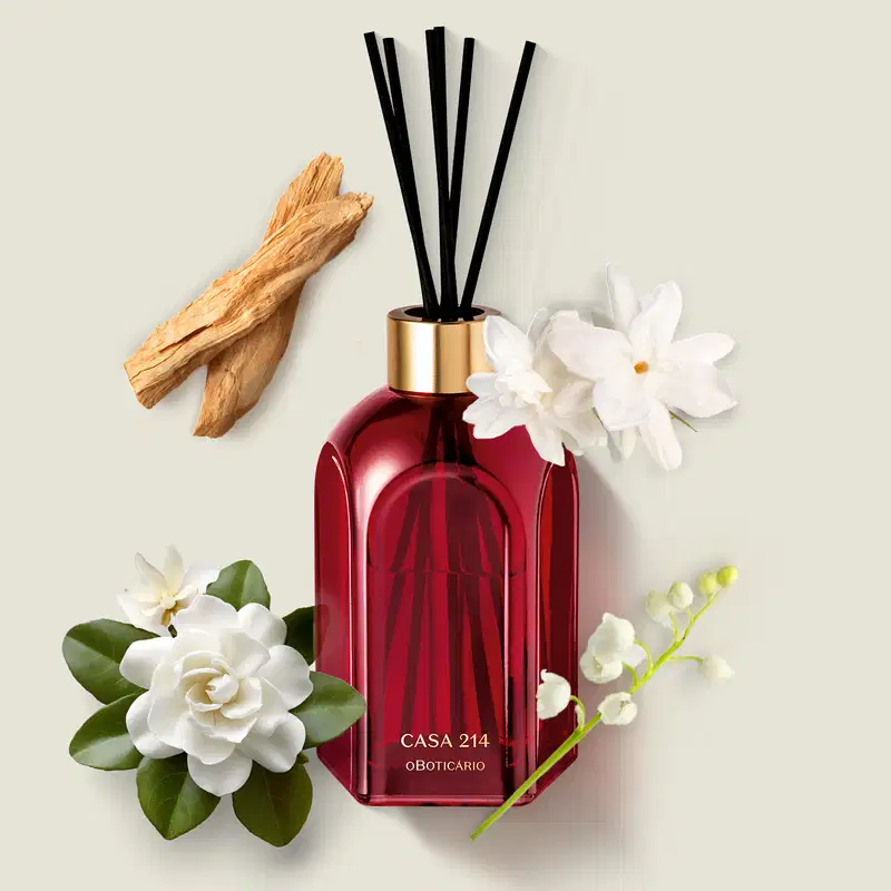 Difusor de ambientes em vidro vermelho com varetas e letras douradas da marca Casa 214 Gardênia Imperial sobre superfície clara com flores brancas e galhos de madeira.