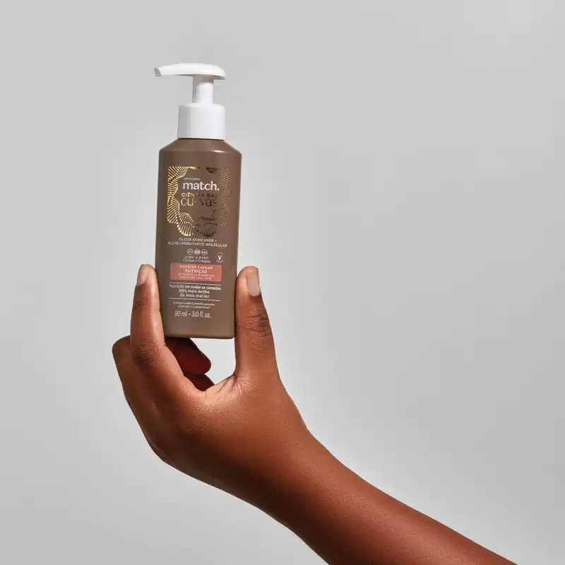 Mão segurando um frasco marrom com bomba dosadora branca e letras brancas e bege claro contendo 90ml de booster capilar Nutrição Match Ciência das Curvas em fundo cinza.
