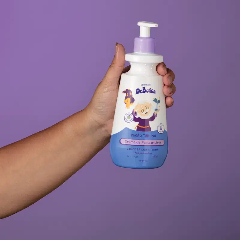 Imagem de uma mão segurando uma embalagem branca e azul com válvula dosadora roxa de creme de pentear infantil para cabelos lisos da marca Dr. Botica em fundo azul escuro.