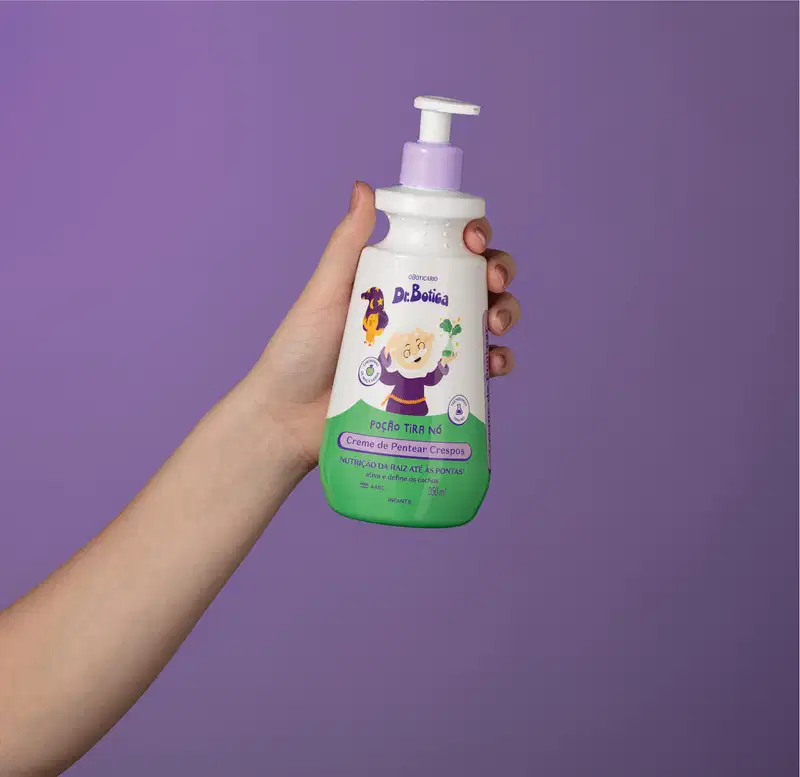 Imagem de uma mão segurando uma embalagem branca e verde com válvula dosadora roxa de creme de pentear infantil para cabelos crespos da marca Dr. Botica em fundo azul escuro.