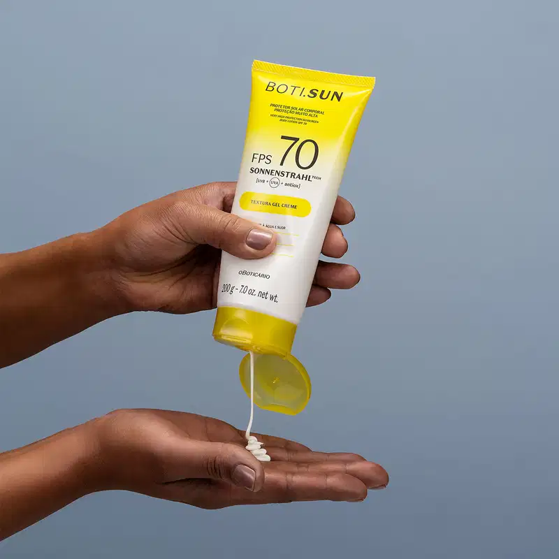 Mão segurando uma Bisnaga amarela e branca com tampa amarela e letras pretas contendo protetor solar corporal FPS 70 da marca Boti.Sun e jogando um pouco do creme branco na outra mão em fundo azul celeste.