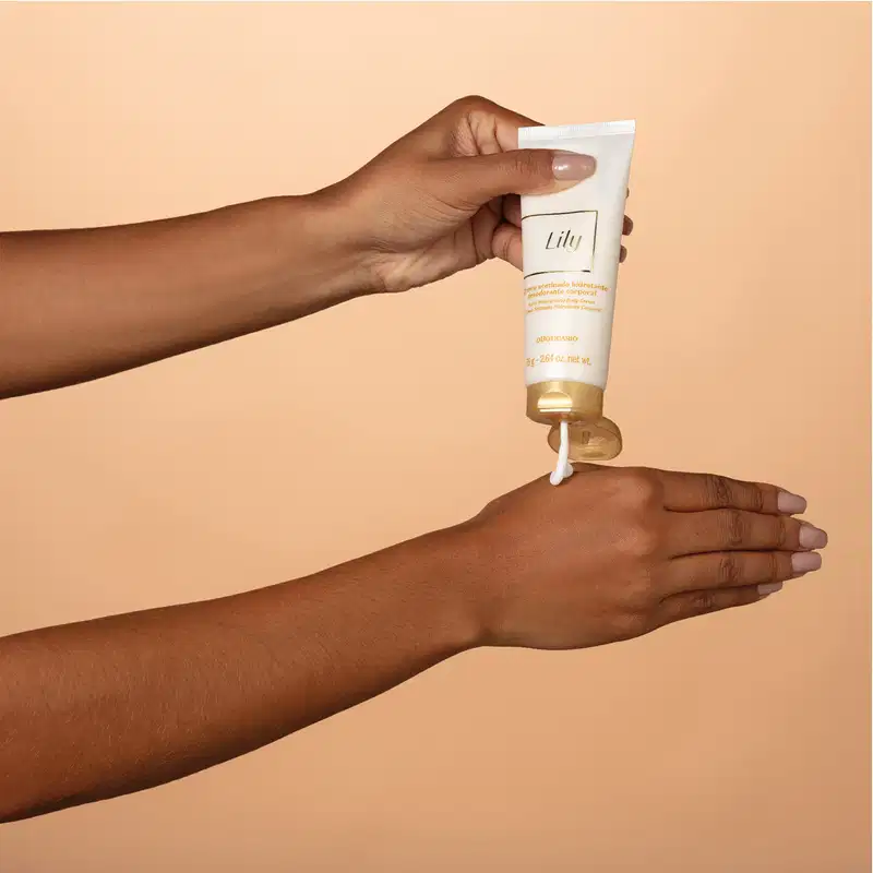 Mão feminina segurando uma bisnaga branca e dourada de creme acetinado hidratante corporal Lily e derrubando um pouco do creme branco na outra mão. O fundo é em tons de bege.