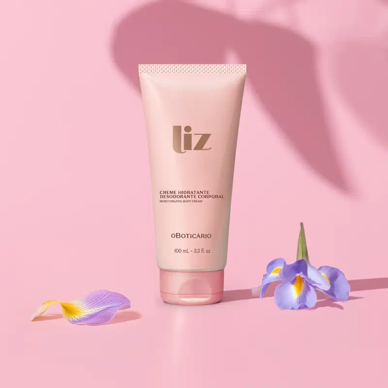 Bisnaga rosa om letras marrons de creme desodorante hidratante corporal da marca Liz em fundo rosa com sombras e flores roxas espalhadas ao redor da embalagem.