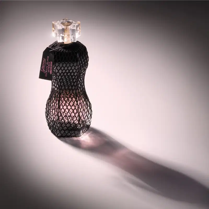 Vidro curvilíneo com renda preta em volta, pulverizador dourado e tampa transparente de desodorante colônia Glamour Secrets Black Intense em fundo claro com a sombra o frasco à frente do item.