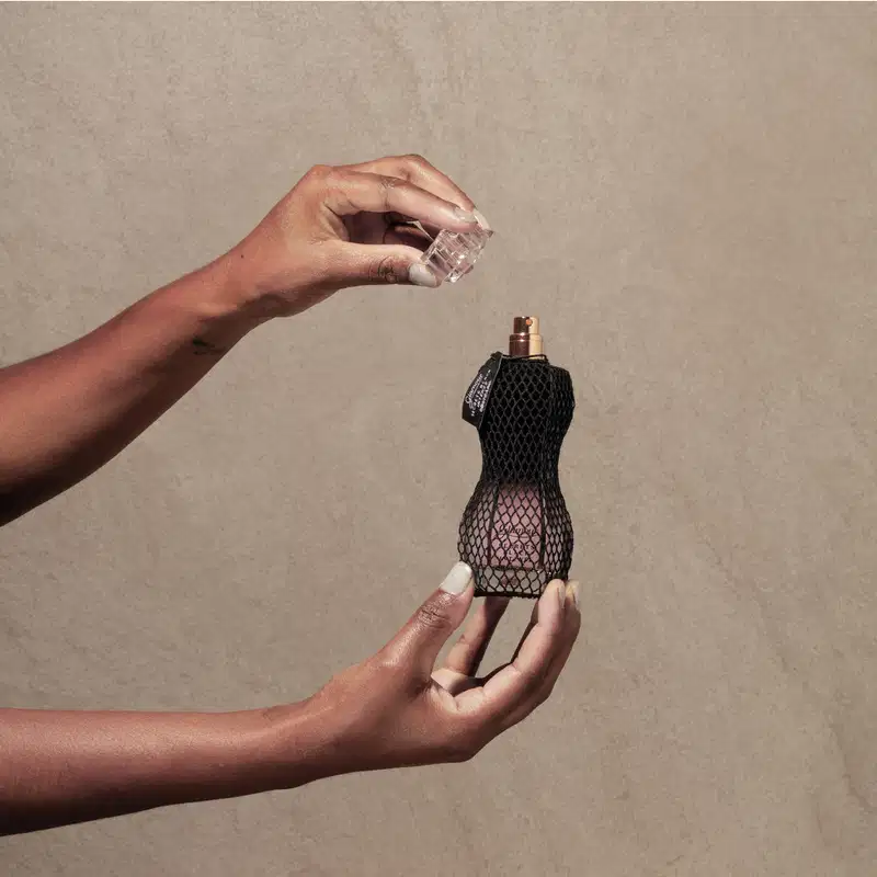 Mão segurando um vidro curvilíneo com renda preta em volta, pulverizador dourado e tampa transparente de desodorante colônia Glamour Secrets Black Intense. A outra mão segura a tampa transparente do produto, tudo em fundo roxo.