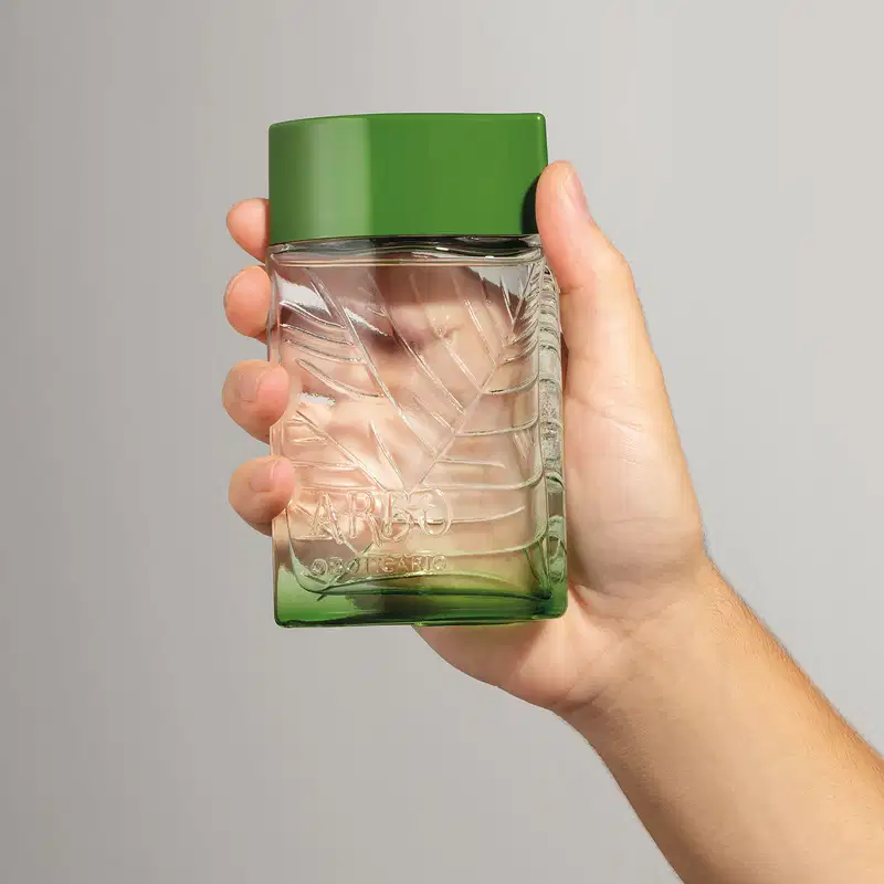 Mão segurando um frasco verde transparente com líquido claro, folhagens em relevo e tampa verde escuro contendo desodorante colônia Arbo Puro em fundo cinza.