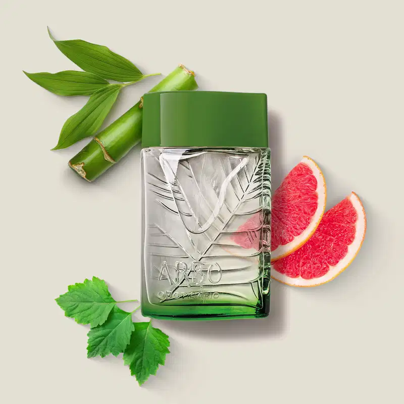 frasco verde transparente com líquido claro, folhagens em relevo e tampa verde escuro contendo desodorante colônia Arbo Puro com folhas verdes e fatias de toranja rosa ao redor, sobre superfície clara.