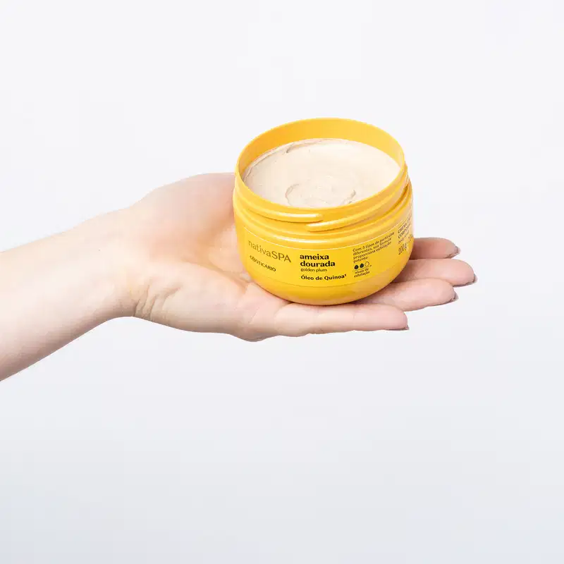 Mão segurando um pote amarelo com tampa e letras pretas contendo creme esfoliante corporal Nativa SPA Ameixa Dourada em fundo azul claro.