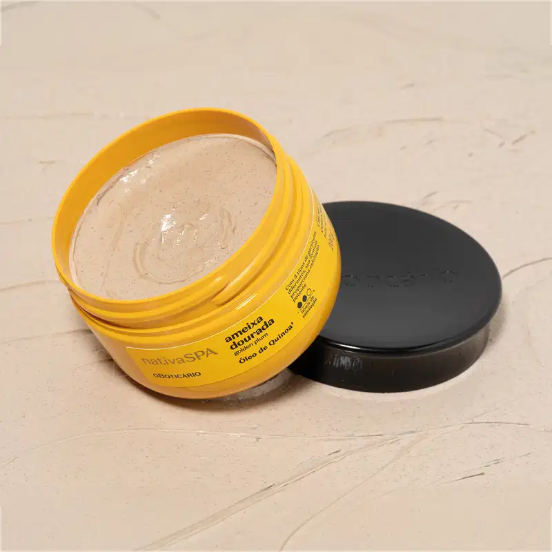 Pote amarelo com letras pretas aberto, mostrando o creme amarelado dentro dele, com a tampa preta ao lado, em superfície bege simulando mármore.