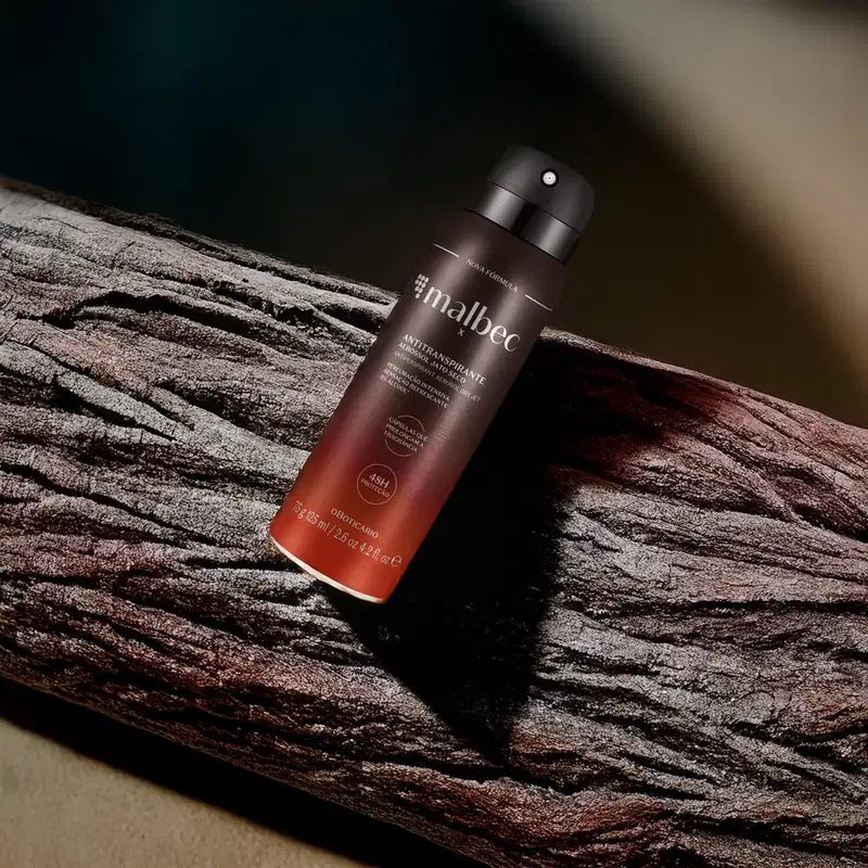 Frasco com degradê de marrom e laranja e letras cinzas de desodorante antitranspirante aerossol da marca Malbec em um fundo escuro e superfície de madeira.