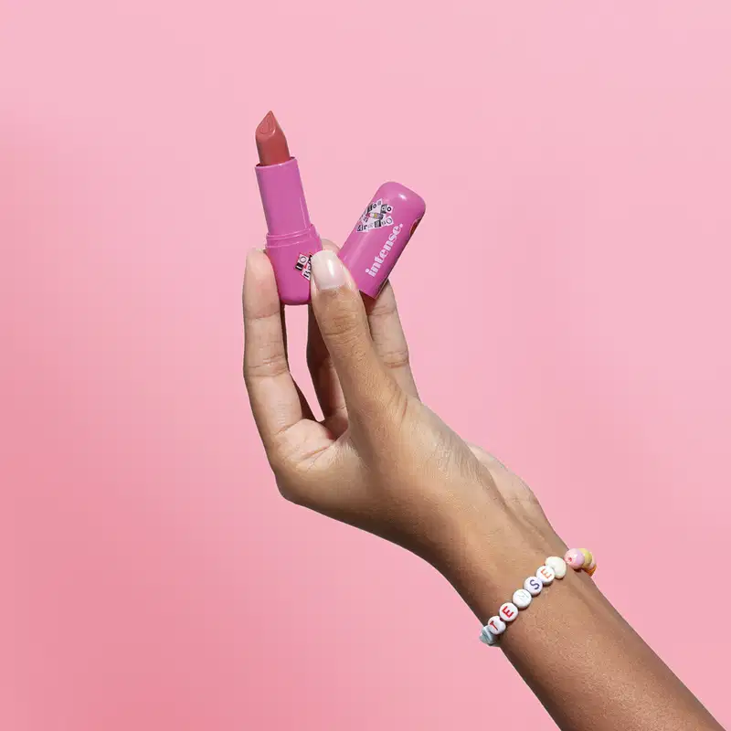 Uma mão feminina segura o batom do Arraso na cor pink rosé da marca Intense em embalagem na cor roxa e a outra mão segura a tampa, na mesma tonalidade, em fundo em tons de rosa.