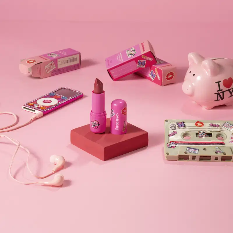 Batom do Arraso na cor pink rosé da marca Intense em embalagem na cor roxa está em cia de uma plataforma rosa com caixa do produto, mp3 player, cofre e fita cassete, tudo em tons de rosa.