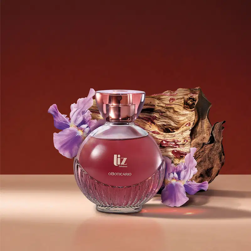 um vidro com líquido vermelho, borrifador dourado, tampa transparente e letras douradas de desodorante colônia Liz Intenso está com muitas flores roxas e madeira ao redor, em superfície rosa e fundo vermelho.