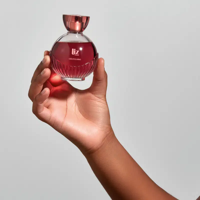 Uma mão está segurando um vidro com líquido vermelho, borrifador dourado, tampa transparente e letras douradas de desodorante colônia Liz Intenso em fundo cinza.