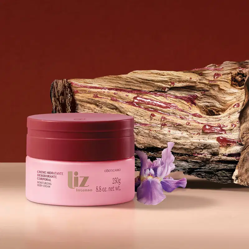 Pote rosa com tampa e letras vermelhas de creme hidratante corporal Liz Intenso ao lado de uma flor roxa e de um galho de árvore, em superfície rosa e fundo vermelho.