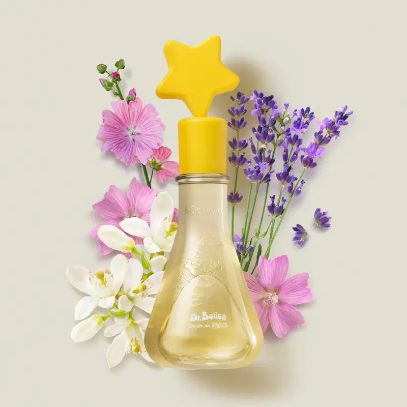 Frasco transparente com líquido amarelo e tampa com estrela na ponta também amarelas de colônia infantil Poção da Amizade da marca Dr. Botica em meio a flores coloridas e decoração amarela sobre superfície clara.