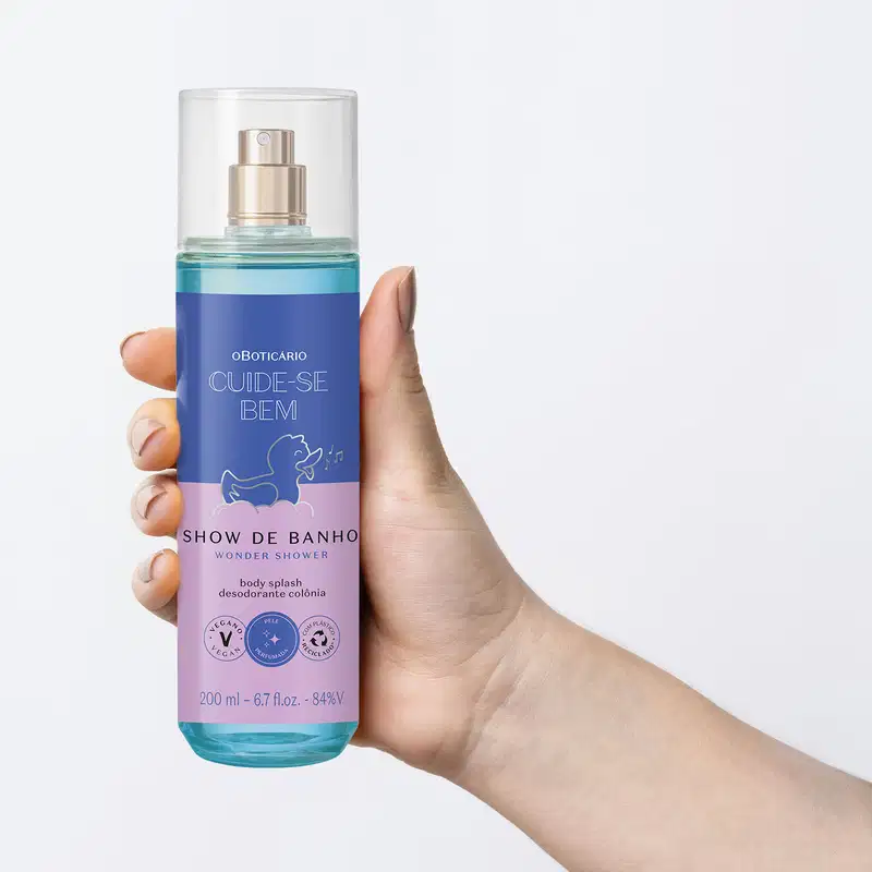 Mão segurando um Frasco com líquido azul, pulverizador prateado, tampa transparente e rótulo azul e lilás com patinho de banho ilustrado de body splash da marca  Cuide-se Bem Show de Banho em fundo azul claro.