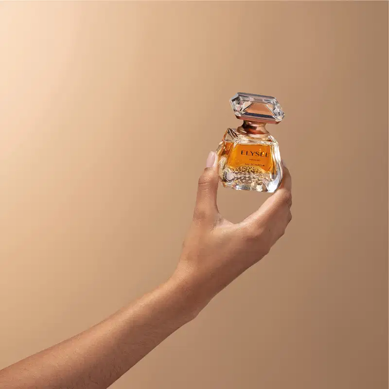 Mão segurando um frasco de Eau De Parfum da marca Elysée em formato geométrico com líquido na cor âmbar e tampa em forma de cristal lapidado, em tons de dourado em fundo com degradê de rosa claro e bege escuro.