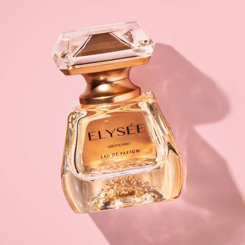 Frasco de Eau De Parfum da marca Elysée em formato geométrico com líquido na cor âmbar e tampa em forma de cristal lapidado, em tons de dourado em fundo rosa claro com sombras.
