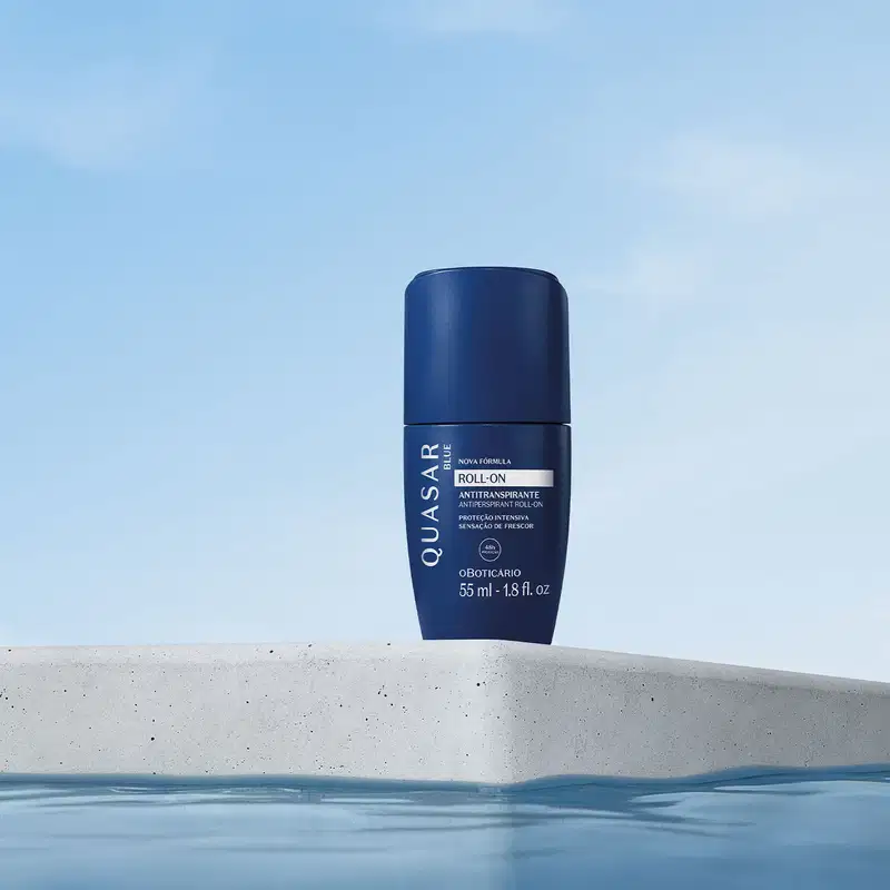 Frasco azul marinho com letras brancas contendo 55ml de antitranspirante desodorante roll-on da marca Quasar Blue flutuando sobre uma superfície de concreto cinza, com céu azul claro ao fundo.