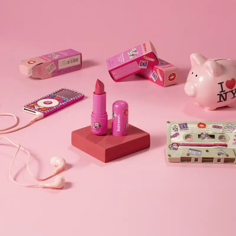 Batom do Arraso Eu Sou Mais  Eu na cor rosa da marca Intense em embalagem na cor roxa está em cia de uma plataforma rosa com caixa do produto, mp3 player, cofre e fita cassete, tudo em tons de rosa.