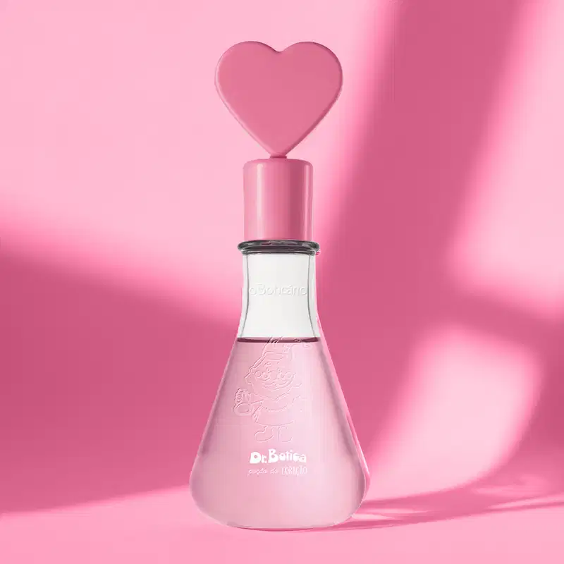 Frasco transparente em formato de vidro laboratorial com líquido rosa, tampa também rosa com coração na ponta e letras brancas no rótulo de colônia infantil Dr. Botica Poção do Coração em fundo rosa com sombras.