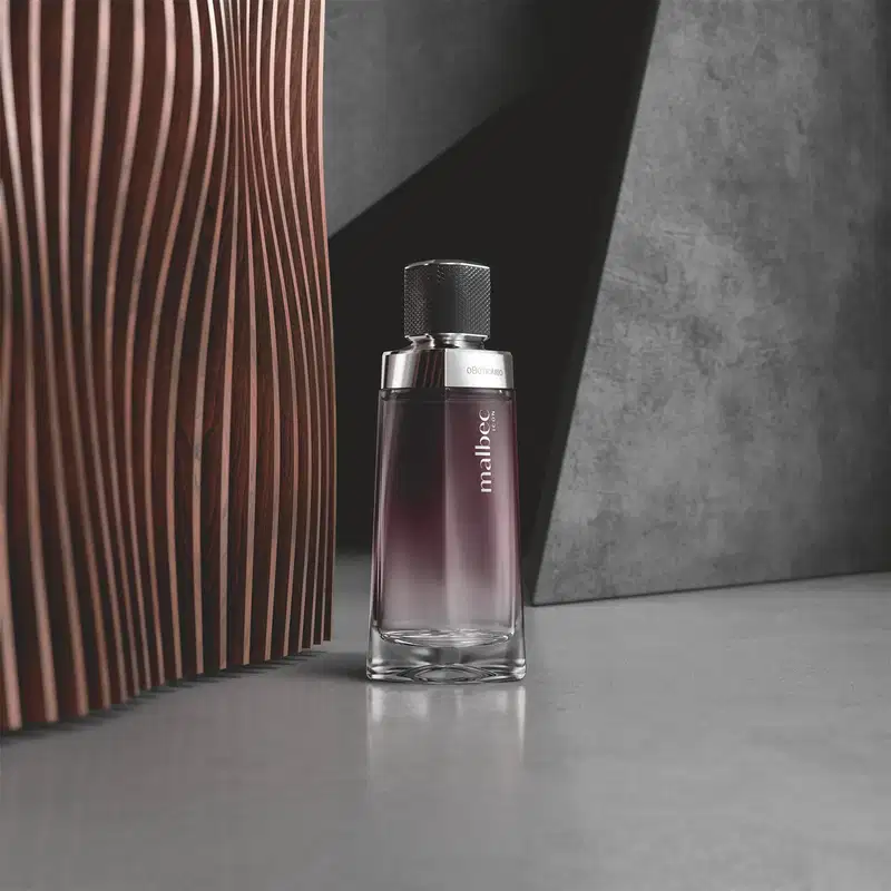 Vidro com degradê de preto e branco, detalhe prateado próximo à tampa preta e rótulo com letras cinza de desodorante colônia Malbec Icon em primeiro plano, sobre superfície cinza, com painel de madeira rosa ao fundo.