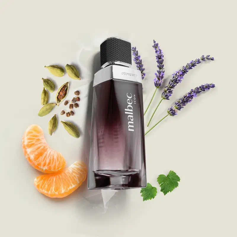 Vidro com degradê de preto e branco, detalhe prateado próximo à tampa preta e rótulo com letras cinza de desodorante colônia Malbec Icon em primeiro plano, rodeado por especiarias, flores de lavanda e fatias de laranja sobre superfície clara.