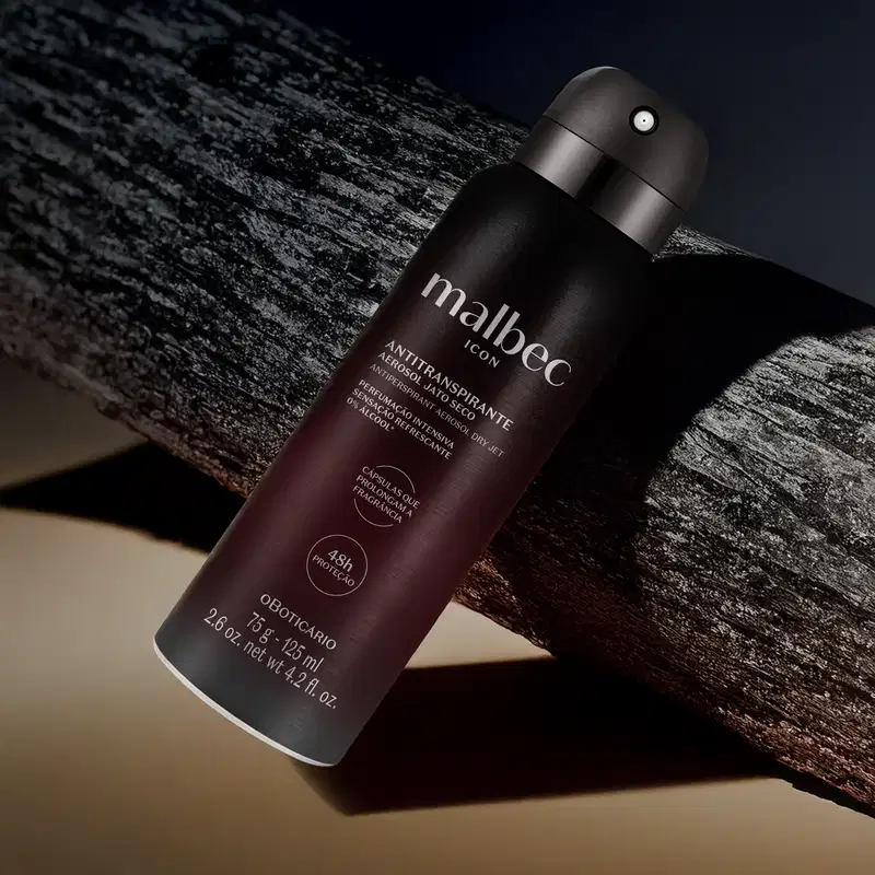 Frasco preto fosco com letras cinzas de antitranspirante aerossol jato seco da marca Malbec Icon sobre superfície de madeira escura, com fundo desfocado e iluminação dramática.