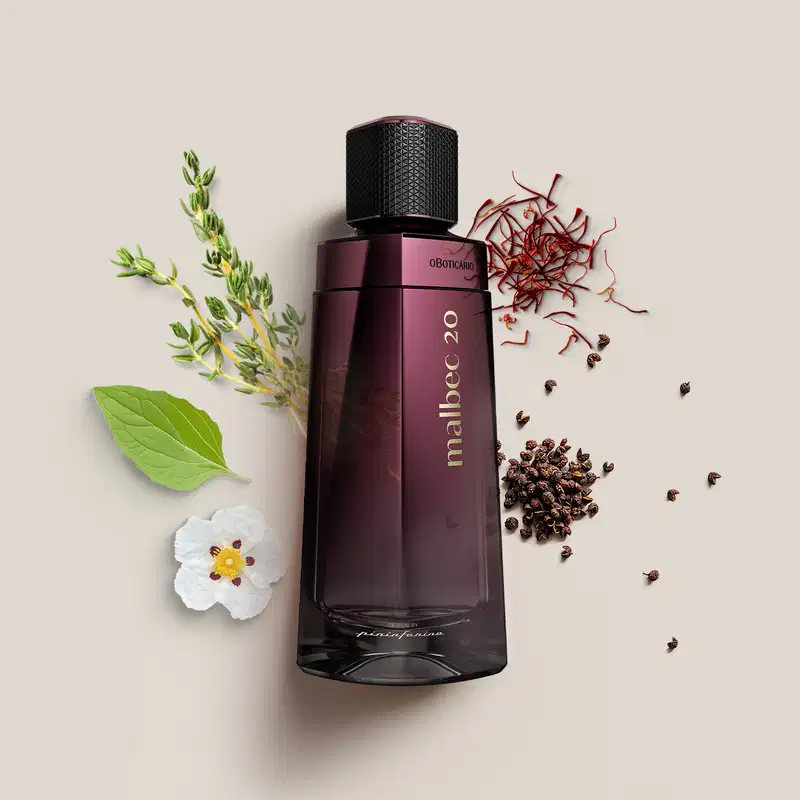Perfume Malbec 20 em frasco retangular de vidro roxo escuro. Embalagem preta texturizada na parte superior. Nome "Malbec 20" e "Oboticário" em letras douradas. Ao redor do frasco, há uma flor branca, folhas verdes, galhos e grãos de pimenta preta.