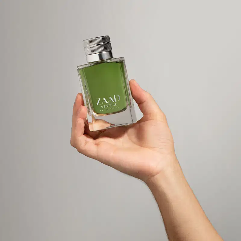 Mão segurando um  vidro retangular na cor verde oliva com tampa texturizada prateada e letras cinza contendo Eau de Parfum Zaad Venture em fundo cinza.