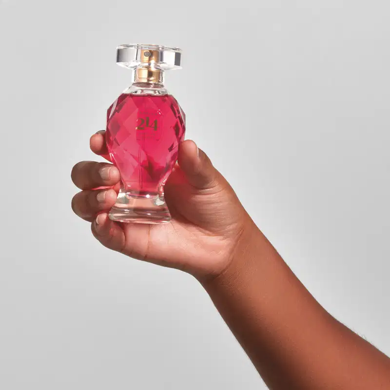 Mão feminina segurando um vidro lapidado em formato de taça com líquido rosa vibrante, tampa transparente, pulverizador e letras douradas contendo 75ml de Eau de Parfum Botica 214 African Sunrise Floral Frutal em fundo azul.