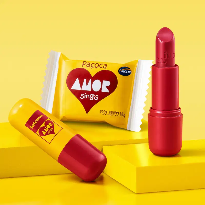 Batom cremoso vermelho da marca Intense By Amor de tampa amarela com tubo e letras vermelhas em cenário amarelo com doce ao redor.