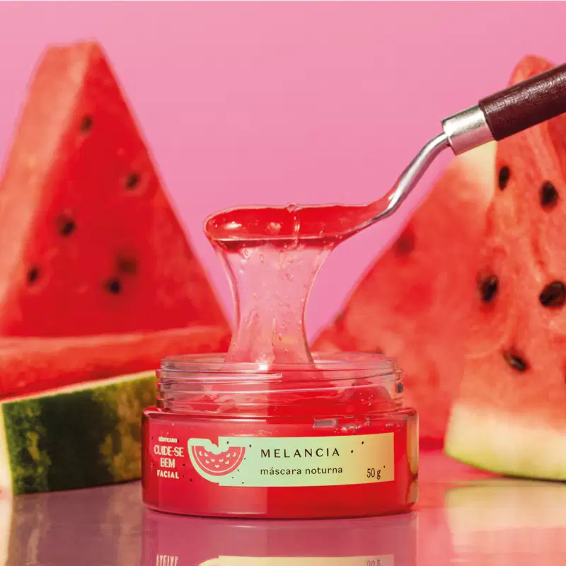 Pote com creme vermelho, tampa verde e rótulo também verde com melancia ilustrada contendo máscara facial noturna Cuide-se Bem Feira Melancia sobre superfície rosa, com fatias de melancia vermelha ao redor e uma colher com um pouco do creme suspensa.
