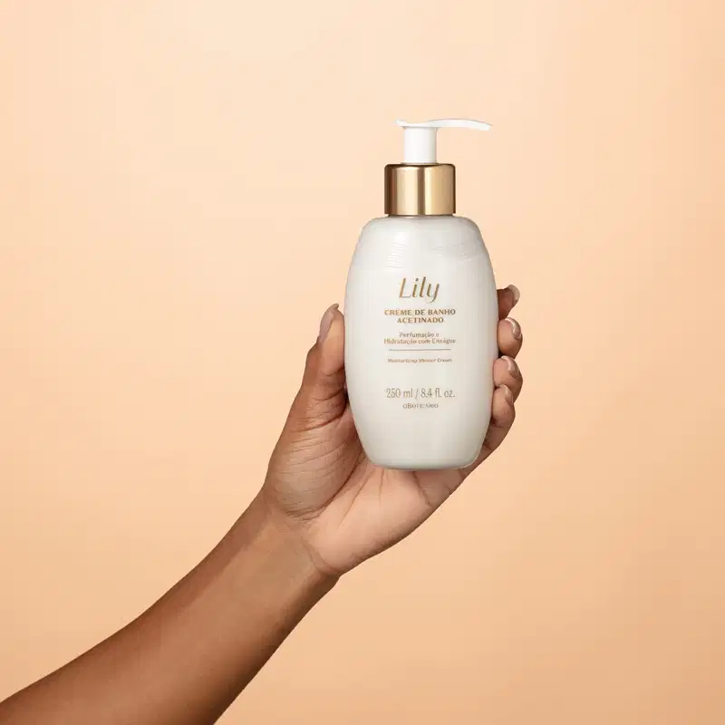 Uma mão feminina está segurando um frasco branco ovalado com detalhe dourado, tampa dosadora branca e letras bege escuras contendo creme de banho acetinado da marca Lily em um fundo em tons de rosa.