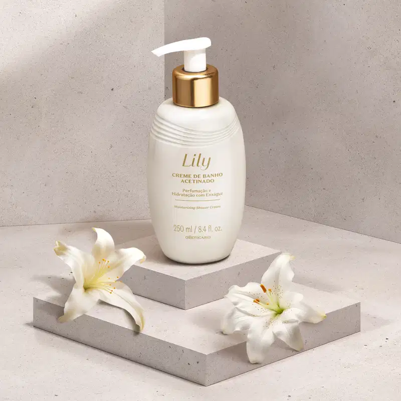 Frasco branco ovalado com detalhe dourado, tampa dosadora branca e letras bege escuras de creme de banho acetinado da marca Lily em plataforma branca com flores brancas ao redor e fundo cinza.