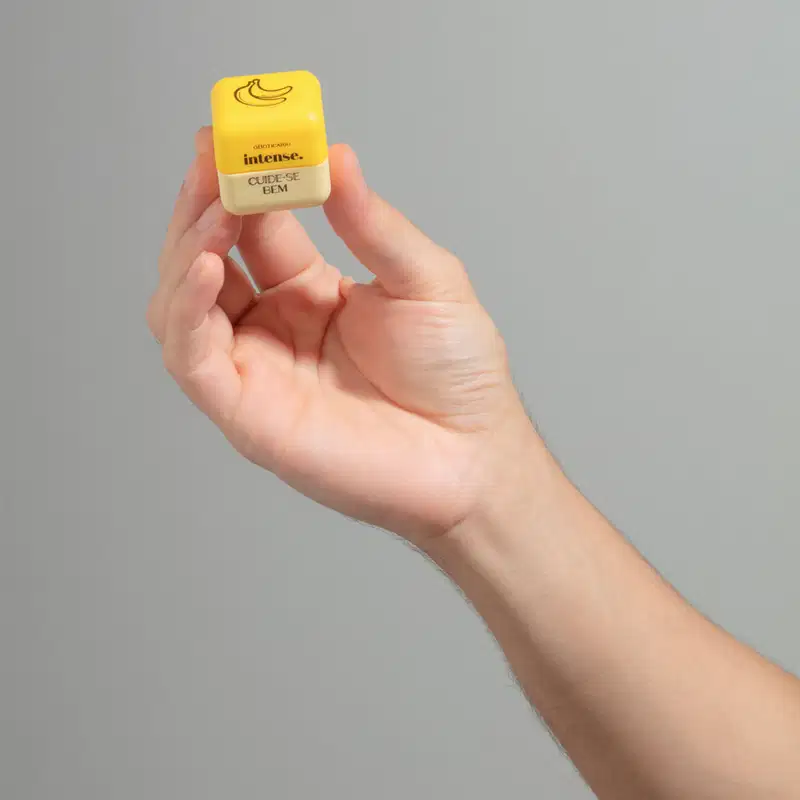 Mão segurando uma embalagem quadrada amarela e branca de balm labial da marca Intense Cuide-se Bem Banana em fundo cinza escuro.