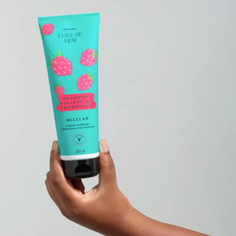 Mão segurando um frasco azul com tampa preta e ilustrações de framboesas na cor rosa contendo shampoo Cuide-se Bem Feira Vinagre de Framboesa em fundo cinza claro.