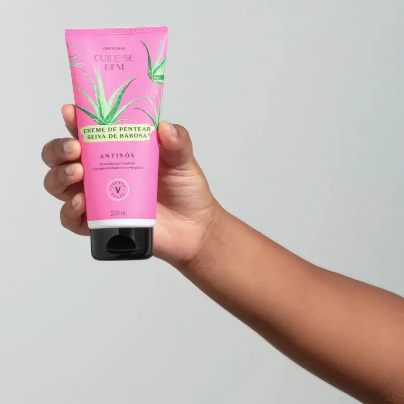 Mão segurando um frasco na cor rosa com tampa preta e ilustração de folhas de babosa contendo creme de pentear Seiva de Babosa da marca Cuide-se Bem Feira em fundo cinza.