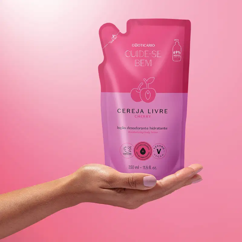 Mão segurando um refil de loção hidratante corporal Cuide-se Bem Cereja Livre em sachê nas cores rosa e roxo com cerejas ilustradas. A imagem está em fundo em tons de rosa.