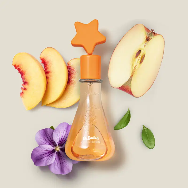 Frasco simulando um vidro laboratorial com líquido laranja e tampa com estrela na ponta também laranja de colônia infantil Poção da Criatividade Dr. Botica. sobre superfície clara, com fatias de maçã, pêssegos  flor roxa e folhas verdes ao redor.
