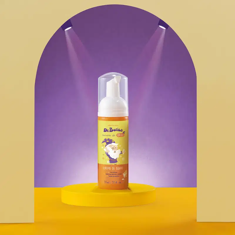 Espuma de banho corporal Nuvem de Ideias Dr. Botica em frasco com tampa branca e rótulo amarelo e laranja com ilustração do personagem da marca em destaque sobre superfície amarela, com iluminação roxa ao fundo, criando um ambiente vibrante e atraente.