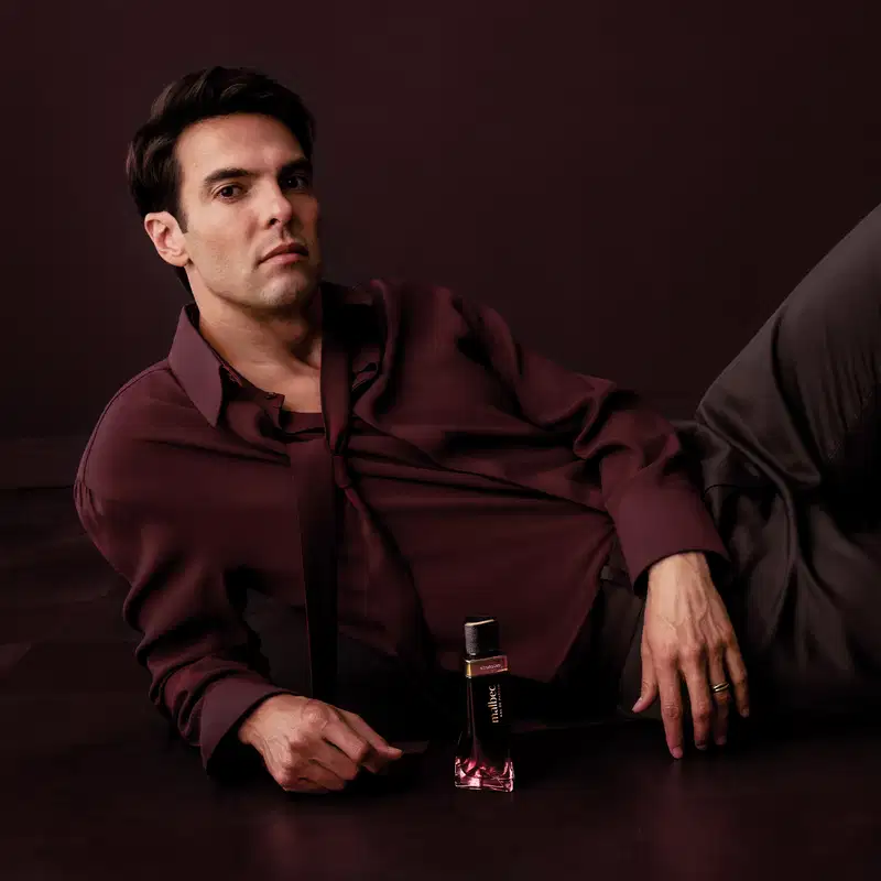 Homem vestido com jaqueta vinho, segurando um item de perfumaria da marca Malbec em ambiente escuro e monocromático.