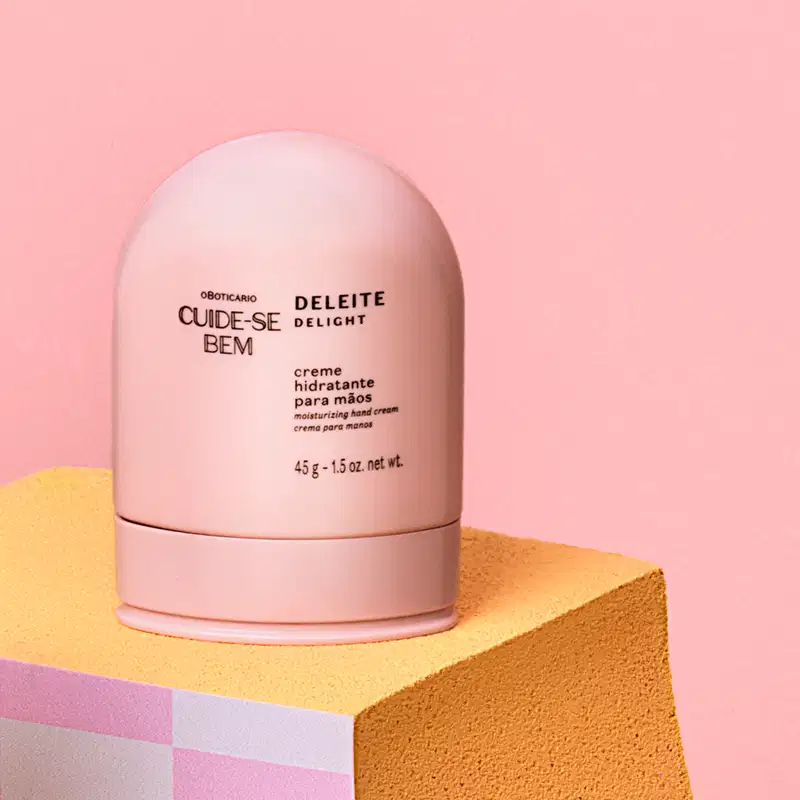 Embalagem de creme hidratante para mãos da marca Cuide-se bem Deleite, em tom rosa claro, sobre superfície amarela com formas geométricas, em ambiente monocromático rosa.