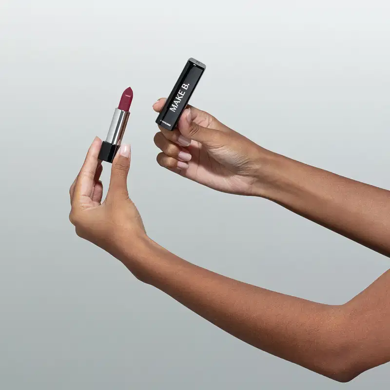 Uma mão feminina está segurando um Batom glossy na cor Vinho Blackbarry da marca Make B. em embalagem prata e preta e a outra mão segura a tampa na cor preta com letras prateadas. O fundo da imagem é cinza.