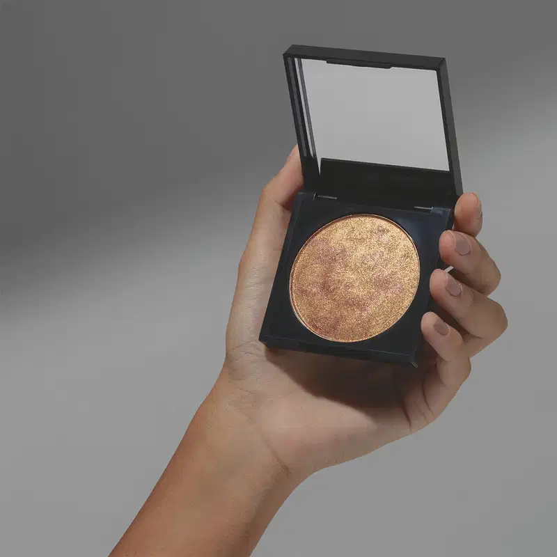 Mão feminina segurando um blush iluminador Marble Shine Gold em tom dourado da marca Make B. em estojo na cor preta e em fundo cinza.